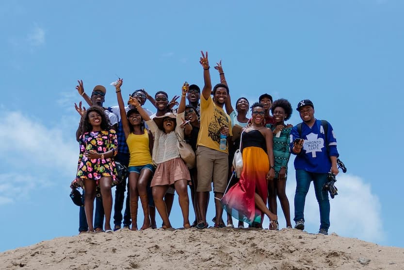 A group of young people standing together on a sandy ridge under a blue sky, laughing and raising their hands.